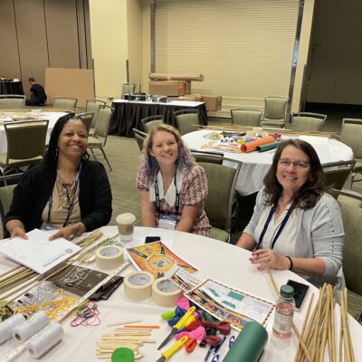 Three participants smile during the Bridging the Divide team building event, seated at a table filled with creative supplies for the bridge-building activity. thumbnail
