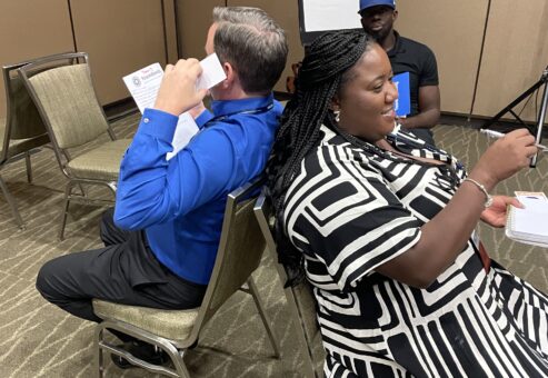 Two participants engage in a collaborative communication exercise, sitting back-to-back as part of the Bridging the Divide team building activity.