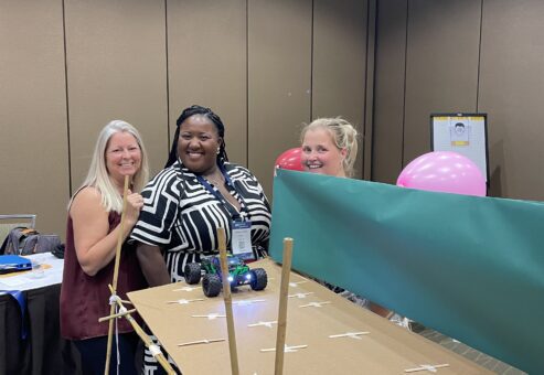 Participants smile next to their completed bridge, ready to test it with a radio-controlled car during the Bridging the Divide team building activity.