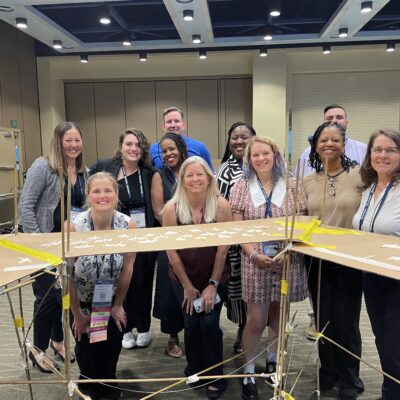 A team poses proudly behind their completed bridge structure during the Bridging the Divide team building activity. thumbnail