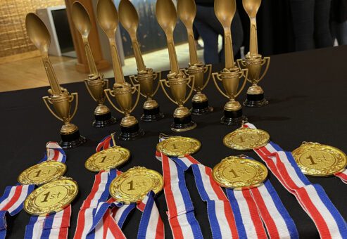 A table displays several gold trophies in the shape of large spoons and medals with '1st' on them. The medals are attached to red, white, and blue ribbons. The trophies and medals are for the culinary team building winners.