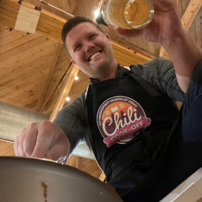 A man is smiling as he participates in a chili cook-off. He is wearing a black apron with the 'Chili Cook-Off' logo prominently displayed. The wooden ceiling and string lights in the background create a cozy atmosphere. The man is holding a glass of beer in one hand and appears to be stirring a pot with the other, fully engaged in the cooking activity. thumbnail