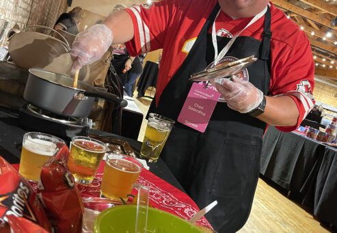 A man wearing a red sports jersey and a black apron is smiling while stirring a pot on a stove during a culinary team building event. He holds a set of tongs in one hand and is wearing plastic gloves. In front of him are cups of beer, a bag of Doritos chips, and a green bowl.