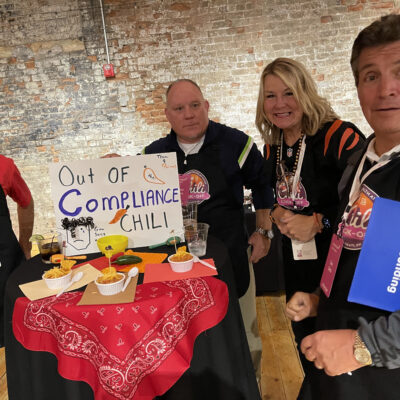Four people standing around a small round table with a red bandana tablecloth. The group is participating in a chili cook-off team building event. The table features a sign that reads 'Out of Compliance Chili' with some illustrations, along with bowls of chili. The group is smiling and wearing aprons with chili cook-off branding. thumbnail