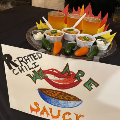 A creatively arranged display featuring small cups of chili garnished with shredded cheese and jalapeños, paired with a dollop of sour cream on the side. The setup includes three glasses of beer placed behind the chili cups, with plastic spoons inserted into each. The presentation is adorned with paper flames, giving a fiery theme to the display. In front, there is a hand-drawn sign that reads 'R-Rated Chili, We Are' alongside a bold illustration of a smiling mouth and a bowl of chili. Several real jalapeño and yellow peppers are placed around the setup. thumbnail