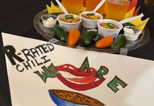 A creatively arranged display featuring small cups of chili garnished with shredded cheese and jalapeños, paired with a dollop of sour cream on the side. The setup includes three glasses of beer placed behind the chili cups, with plastic spoons inserted into each. The presentation is adorned with paper flames, giving a fiery theme to the display. In front, there is a hand-drawn sign that reads 'R-Rated Chili, We Are' alongside a bold illustration of a smiling mouth and a bowl of chili. Several real jalapeño and yellow peppers are placed around the setup.