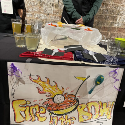 A table at a chili cook-off team building event displaying a colorful sign that reads 'Fire in the Bowl' with a graphic of a chili bowl on fire. The table has various chili-related items, including drinks, cups, and utensils. A person in the background is wearing a black apron with a 'Chili Cook-Off' logo. thumbnail