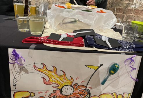 A table at a chili cook-off team building event displaying a colorful sign that reads 'Fire in the Bowl' with a graphic of a chili bowl on fire. The table has various chili-related items, including drinks, cups, and utensils. A person in the background is wearing a black apron with a 'Chili Cook-Off' logo.
