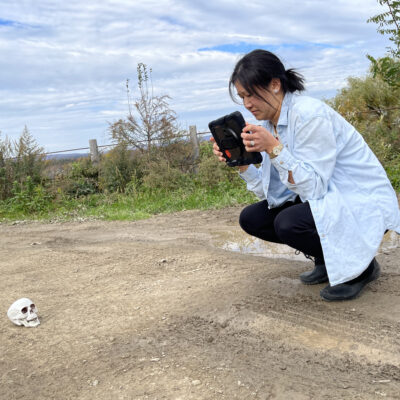 A woman crouches down on a path, using a tablet to photograph a skull placed on the ground, simulating a crime scene investigation as part of a team building activity focused on making a movie. thumbnail
