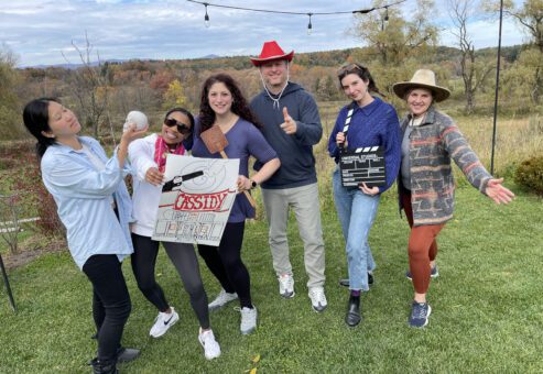 A group of six people pose outdoors, holding various props like a film clapperboard, a poster, and a red cowboy hat, participating in a creative team building activity themed around film production and storytelling.