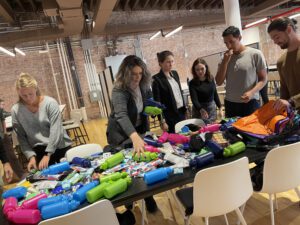 Participants gather around a table filled with supplies as they work together to assemble care kits during the Helping Hands team building event. The group organizes items such as water bottles, hygiene products, and snacks for community members in need.