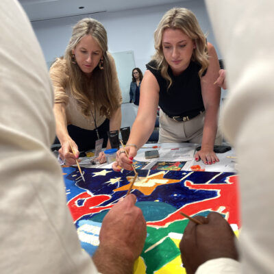 Two team members concentrate as they add final touches to their section of a vibrant mural during The Big Picture team building event, fostering collaboration and creativity. thumbnail
