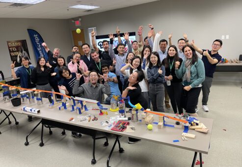 A group of excited participants celebrate their collaborative success after completing a Rube Goldberg-inspired contraption during the Domino Effect team building event.