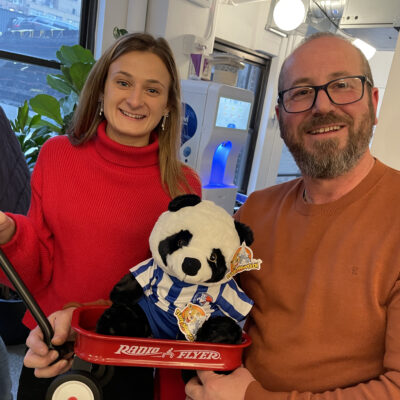 A man and a woman stand together smiling, holding a small red Radio Flyer wagon with a stuffed panda bear dressed in a blue and white striped outfit. They are participating in a charitable team building event, showing their completed project with pride and joy. thumbnail