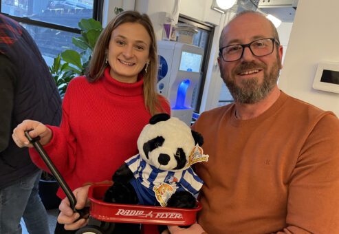 A man and a woman stand together smiling, holding a small red Radio Flyer wagon with a stuffed panda bear dressed in a blue and white striped outfit. They are participating in a charitable team building event, showing their completed project with pride and joy.