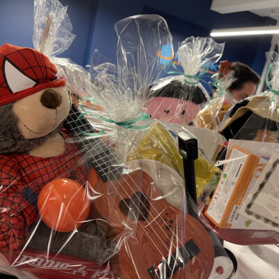 A close-up image of a red Radio Flyer wagon filled with a gift set for charity. The wagon includes a stuffed teddy bear dressed in a superhero outfit, a small ukulele, a maraca, and various art supplies. The entire wagon is wrapped in clear gift wrap, ready for donation at the Toys for Tykes ‘Arts & Music’ team building event. thumbnail