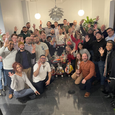 A large group of people smiling and waving for a group photo. They are gathered around a collection of decorated toys and gifts, assembled during a CSR team building program. thumbnail