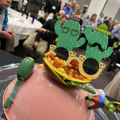A festive salsa presentation featuring a bowl of fresh salsa garnished with playful cactus-shaped glasses, maracas, and a mini piñata, capturing the lively spirit of the Salsa Showdown team building event. thumbnail