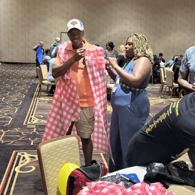 A man wearing a baseball cap is being fitted with a checkered red and white fabric draped over his shoulders, resembling a makeshift outfit. He is being assisted by a woman standing next to him, who is holding part of the fabric as she adjusts the look. They are participating in a fun team building activity in a large conference room with other groups in the background working on similar projects. thumbnail