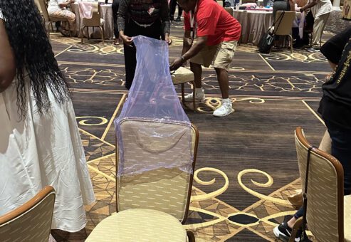 Two participants in a team building activity are working together to cover a chair with a transparent purple material. The man is securing the fabric at the bottom, while the woman stretches the material across the back of the chair.