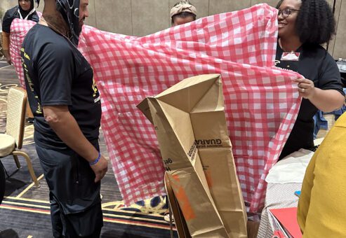 Two participants at a team building event stand, with one holding a large red and white checkered fabric, while the other appears to be modeling it or assisting in its setup. Cardboard and other recyclable materials are visible, being used to create an outfit.