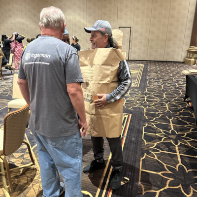 Two participants are engaged in conversation during a team building event. The man on the right is wearing an outfit made from a large brown paper bag, styled as a tunic. Both individuals appear to be enjoying the activity, and the event is clearly focused on fashion, creativity and fun. thumbnail