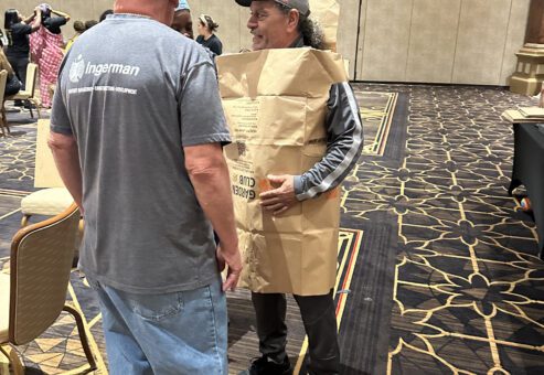 Two participants are engaged in conversation during a team building event. The man on the right is wearing an outfit made from a large brown paper bag, styled as a tunic. Both individuals appear to be enjoying the activity, and the event is clearly focused on fashion, creativity and fun.