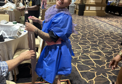 A smiling participant wearing a creatively made outfit composed of colorful recyclable materials, standing in a ballroom. The person is holding a piece of material while others in the background work on their designs. It is a team building event themed around fashion design, with participants using unconventional materials to create outfits.