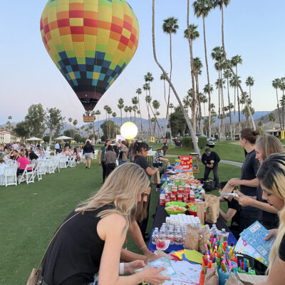 Outdoor corporate charitable team building event with a colorful hot air balloon in the background and participants decorating kits at a table filled with supplies. thumbnail