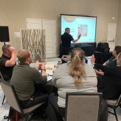 Participants attentively listen to a facilitator explaining striking techniques during a board-breaking team building session focused on overcoming obstacles and personal growth. thumbnail