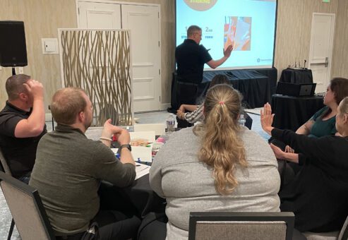 Participants attentively listen to a facilitator explaining striking techniques during a board-breaking team building session focused on overcoming obstacles and personal growth.