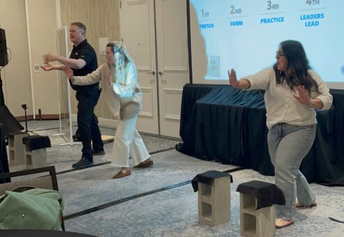 Two participants practice board-breaking techniques on stage during a team building event, guided by a facilitator.