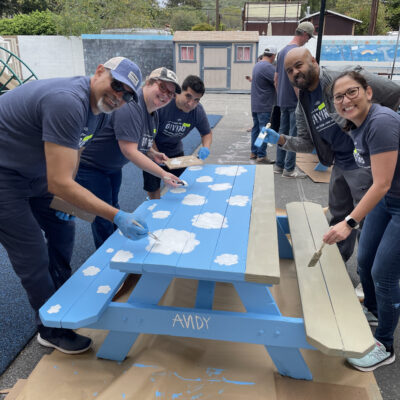 A group of five volunteers working together to paint an outdoor picnic table at a charitable team building event. The table is painted in bright blue with white clouds. All participants, wearing matching grey shirts and blue gloves, are smiling as they contribute to the project. thumbnail