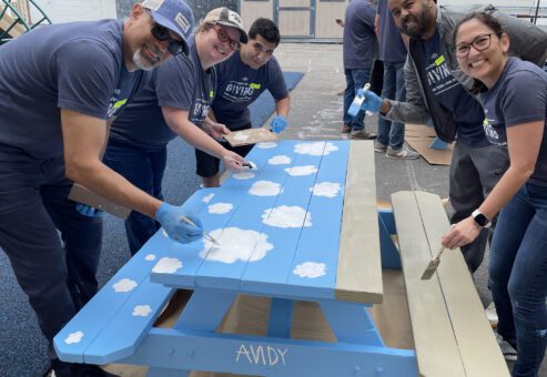 A group of five volunteers working together to paint an outdoor picnic table at a charitable team building event. The table is painted in bright blue with white clouds. All participants, wearing matching grey shirts and blue gloves, are smiling as they contribute to the project.