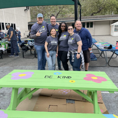 A group of six people standing together, smiling in front of a brightly painted picnic table that reads 