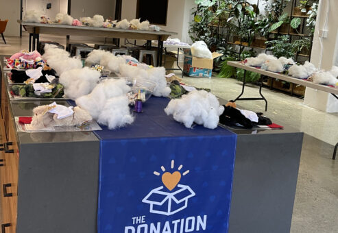 Donation Station setup with tables full of kit materials, including white packing material and small items for assembling donation kits. A banner with 'The Donation Station' is displayed in a brightly lit room, ready for a corporate charitable team building event.