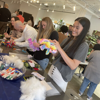 Participants at a Donation Station event assembling colorful donation kits, with supplies like markers and stuffing on the table. A woman in the foreground smiles as she works on a stuffed toy. thumbnail