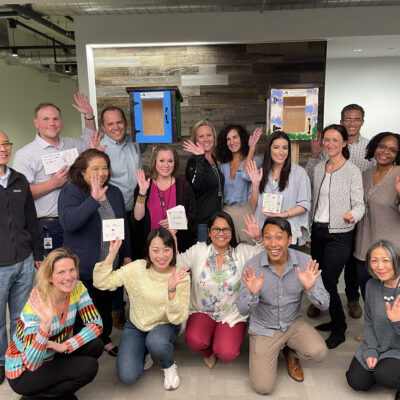 Group of participants smiling and waving in front of two completed Little Free Libraries during the Little Team Library team building event. thumbnail