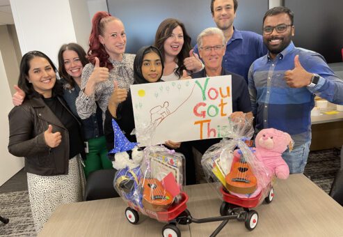A group of eight people stand together smiling and giving thumbs up around two small Radio Flyer wagons filled with teddy bears and toys. One person is holding a handmade sign that says 'You Got This!' with a colorful drawing. The team appears happy and proud, showcasing their contributions during a team building activity focused on charity and giving back.