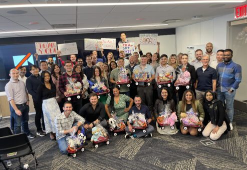A large group of people smiling and posing together in a modern office space. They are holding and sitting with small red Radio Flyer wagons filled with various toys, teddy bears, and musical instruments, all wrapped in gift wrap. Behind them, some participants are holding up hand-drawn signs with motivational messages like 'You Got This!' This photo captures the conclusion of the Toys for Tykes ‘Arts & Music’ CSR team building event.