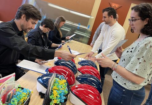 Participants in a team building event stand at a table with red and blue bicycle helmets, discussing instructions and checking documents. Gift baskets with colorful decorations are also displayed, highlighting the preparation phase of a Charity Bike Build event.