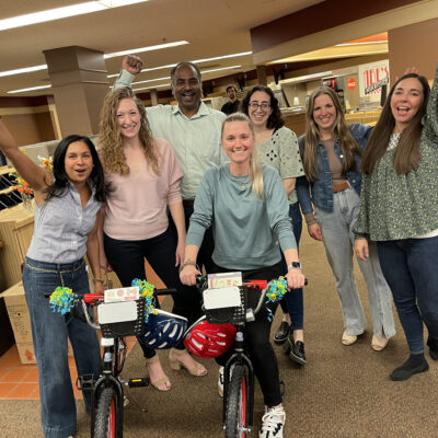 A cheerful group of seven team members celebrates their successful Charity Bike Build. Two decorated bikes stand in front of them, with the group raising their arms in victory. The atmosphere in the room is lively and filled with camaraderie. thumbnail