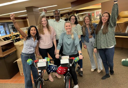 A cheerful group of seven team members celebrates their successful Charity Bike Build. Two decorated bikes stand in front of them, with the group raising their arms in victory. The atmosphere in the room is lively and filled with camaraderie.