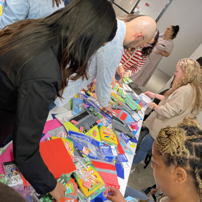 Participants at a Tools for Schools CSR event gather around a table filled with school supplies as they pack colorful folders, notebooks, and other materials into backpacks to give to students. thumbnail