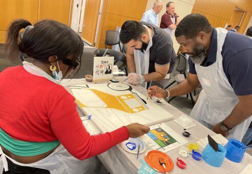 Team members collaborate to paint a section of a large mural during The Big Picture team building event, working together to create a unified masterpiece.