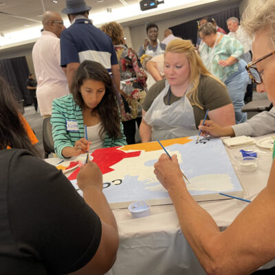 A group of participants sits around a table during The Big Picture team building event, painting a canvas section with various colors. They focus on filling in their assigned part of the mural, collaborating to complete the larger artwork. thumbnail