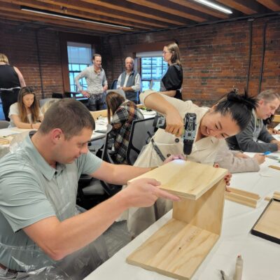 Enthusiastic participants work together to assemble a Little Free Library during a team building event. One team member uses a power drill while another holds the wooden pieces steady, demonstrating teamwork, coordination, and problem-solving as they build the library structure. thumbnail