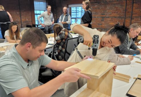 Enthusiastic participants work together to assemble a Little Free Library during a team building event. One team member uses a power drill while another holds the wooden pieces steady, demonstrating teamwork, coordination, and problem-solving as they build the library structure.