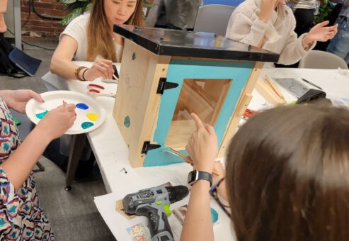 Participants focus on painting a Little Free Library door with bright orange during a collaborative team building event. The hands-on activity fosters creativity and teamwork as teams work together to build and decorate the library for community use.