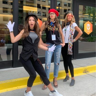 Three women dressed in creative costumes, including hats and gloves, posing energetically outside a venue during a Corporate Film Festival team building event, showcasing their excitement and team spirit. thumbnail